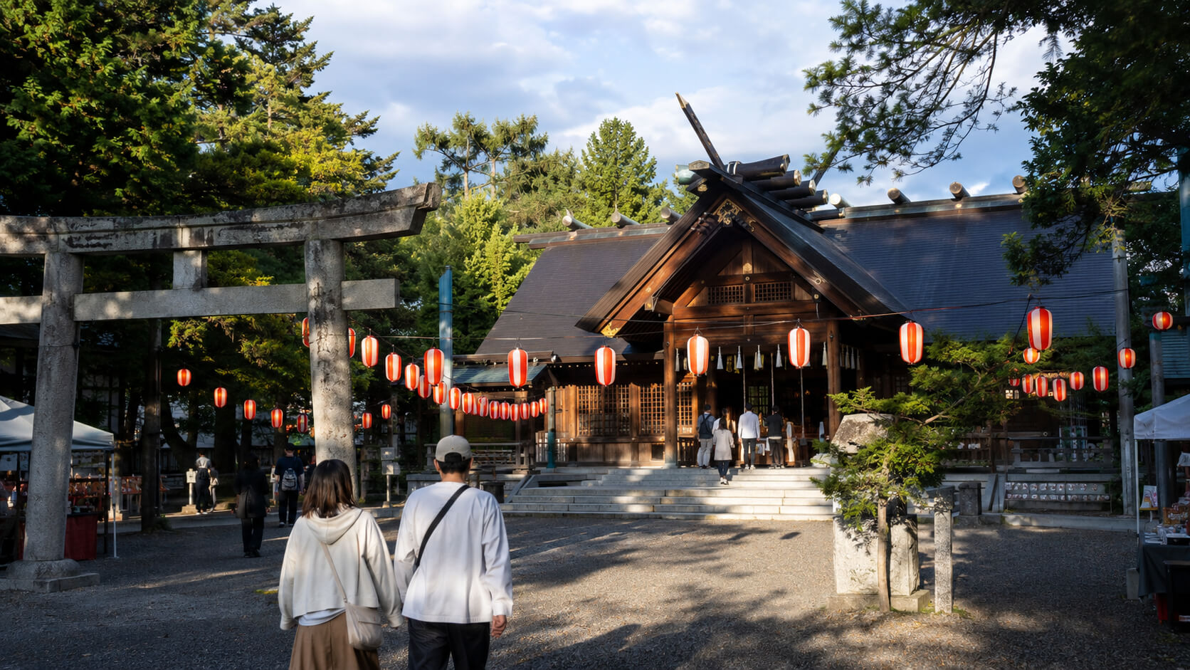 北海道札幌の小さな神社の秋祭り、夕暮れの境内に提灯が灯り始める情景。札幌軟石風の石鳥居と木造社殿を中心にした実写写真風。参拝客が自然に行き交い、地域密着のお祭りらしい温かさを感じる構図。文字なし、ロゴなし、16:9。